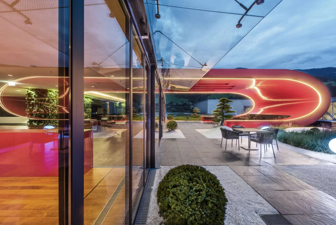 Glasfassade mit Vordach, dahinter Dachterrasse mit beleuchteter roter Loungeschleife in der Dämmerung